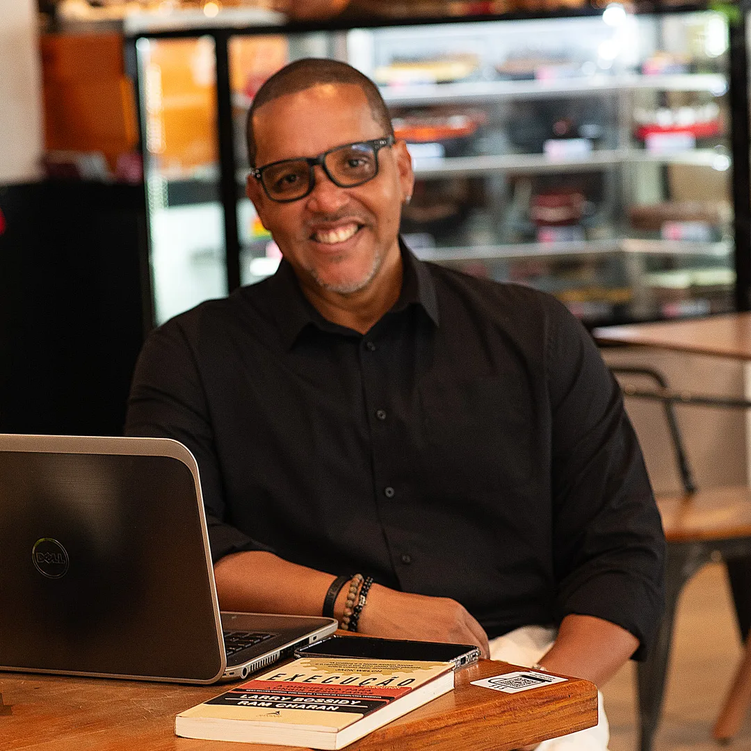 Luetil Neto, fundador da FONS Consultoria, sorrindo, de camisa preta, sentado à mesa de um café com notebook e livro